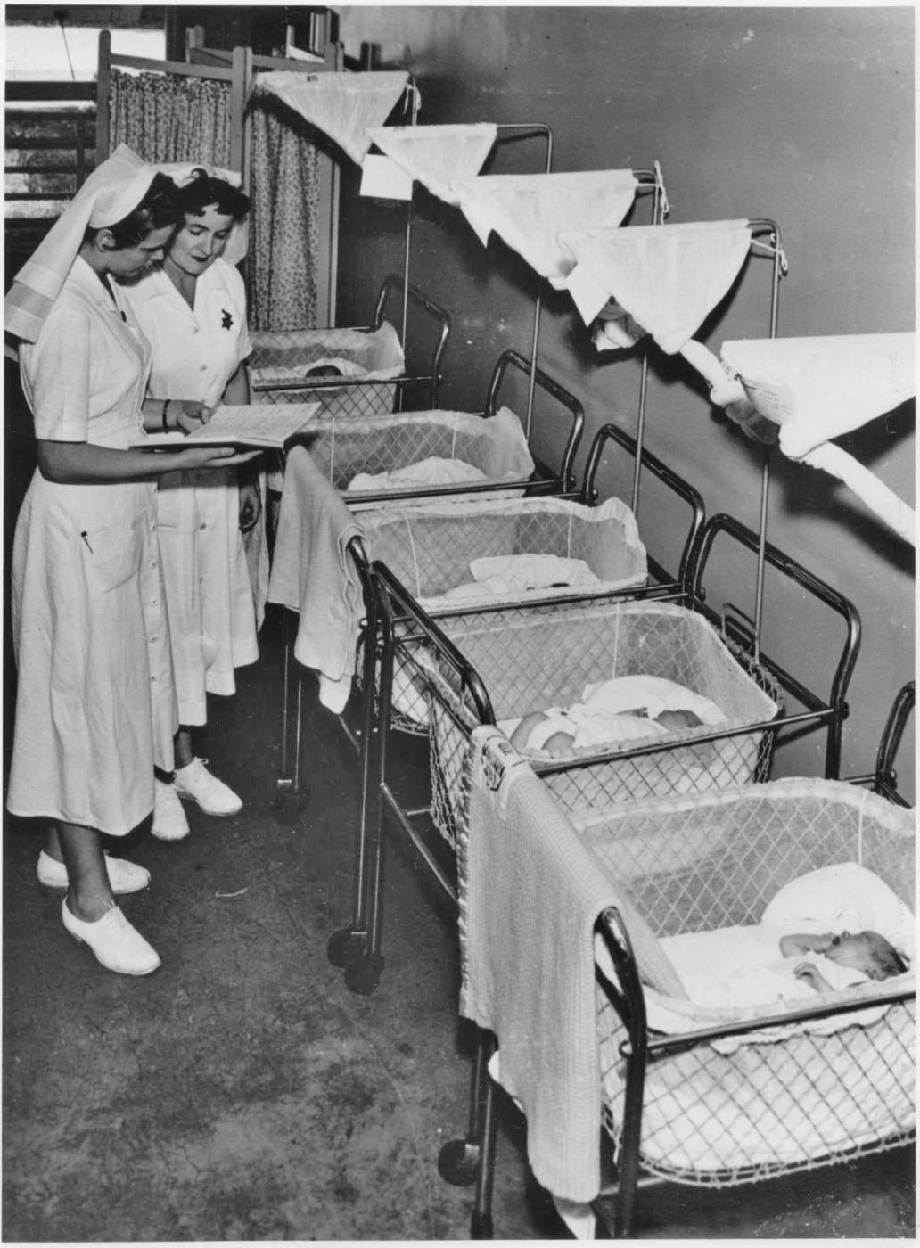Image of two nurses looking at a book while standing in front of 5 bassinets