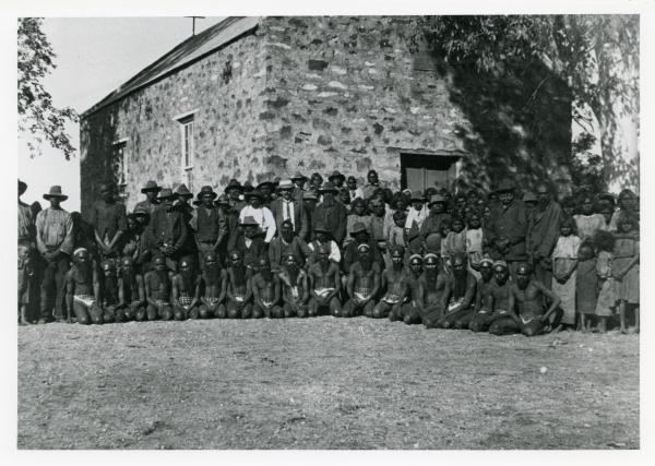Depicts a large group of Aboriginal people in front of a large stone building.